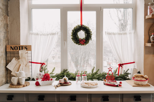 Christmas Kitchen Interior Decorated Xmas Wreath, Pine Tree Branch near Window. Utensil, Flatware for Tea, Decorations, Cookies and Bread on Wooden Table. Desk with Paper Lists Notes on Grunge Wall