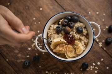 Vegan breakfast, woman's hand and porridge top view on wooden table with oat flakes, banana, blueberries, chia, cinnamon, maple syrup and strawberry jam. Horizontal.