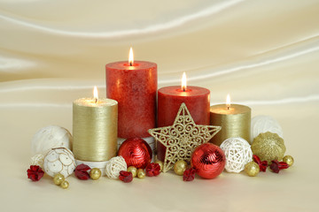 Fototapeta na wymiar Red and gold burning Christmas candles surrounded by natural potpourri elements and gold stars on a cream satin draped background