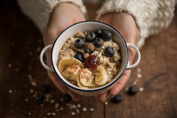 Woman hands offering a healthy snack, porridge with oat flakes, banana, blueberries, chia, cinnamon, maple syrup and strawberry jam. Horizontal. Top view.