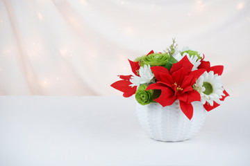 Christmas table floral arrangement with red poinsettia flowers, green roses and white daisies on a white ceramic pot with a white draped fabric background.