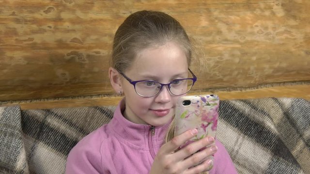 Serious girl reads book on smartphone and smiles.On the sofa covered with a warm woolen blanket, a little teenage girl in glasses sits and reads. Close-up portrait with a camera offset.