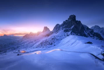 Fototapeten Schneebedeckte Berge und unscharfe Autoscheinwerfer auf der kurvenreichen Straße nachts im Winter. Wunderschöne Landschaft mit schneebedeckten Felsen, Haus, Bergfahrbahn, blauer Sternenhimmel bei Sonnenuntergang in den Dolomiten, Italien © den-belitsky