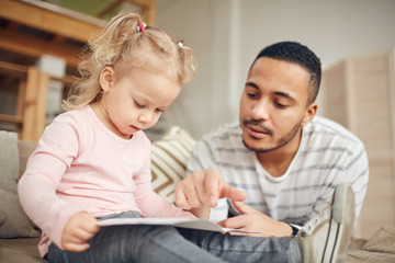 Portrait of young stay at home dad reading books with cute little daughter while enjoying tine together in home interior