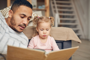 Portrait of young man reading book to cute little girl while enjoying time together at home, copy...