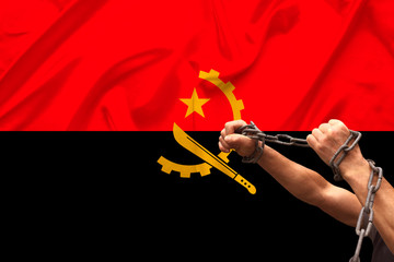 male hands of a prisoner in iron chains against the background of the national flag of Angola on...