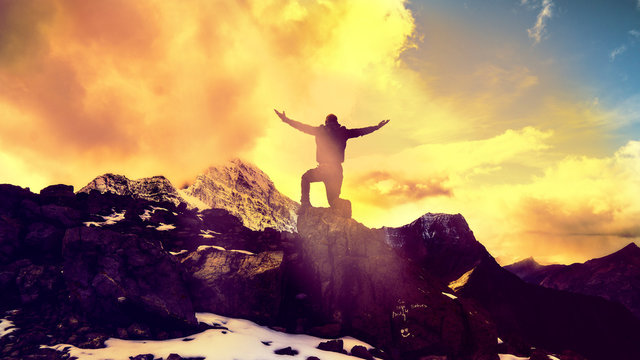 Man Praying Kneeling With Arms Open On Epic Mountain Top Summit With Light Shining With Arms Out On Top Of Mountain Peak