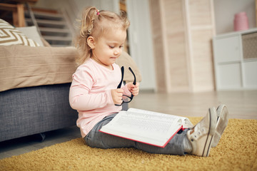 Full length portrait of cute little girl reading big book and plying with glasses while sitting on floor in cozy home interior, copy space