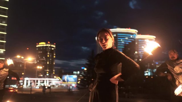 Happy female dancer moves with glowing torches with group of fireshow artists. They present synchronously choreography while the camera goes around them. Slow motion, city landscape.