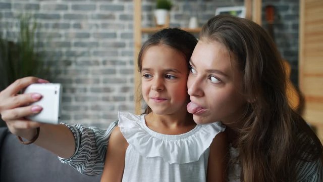 Playful young mother is taking selfie with adorable little daughter having fun in house enjoying funny acitivity. People, positive emotions and motherhood concept.