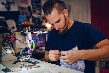 Adult man tailor with beard sitting by his sewing machine at his work shop making a bag checking stitches handmade creative craft working at home