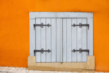 A small white window is closed by wooden shutters with decorative hinges on a bright orange wall.