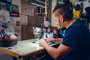 Adult man tailor with beard sitting by his sewing machine at his work shop making a bag checking...
