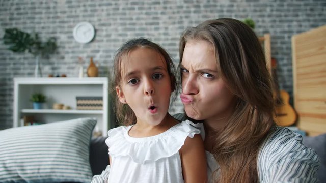 POV of funny young woman and cute girl mother and daughter making funny faces taking selfie at home enjoying funny activity. Youth, photos and emotions concept.