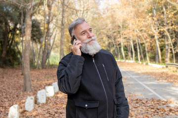 old man with a quiet beard talking on the smartphone on a road with leaves