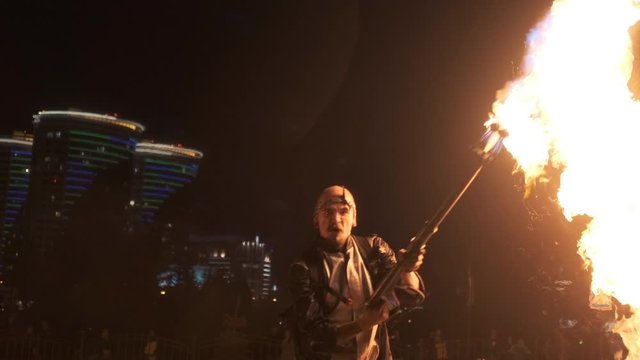 Aggressive brutal showman demonstrate strongness with flamethrower. Rush and stream of fire, fuel spray. Fireshow with flame cannon or machine gun. Night city. Slow motion.