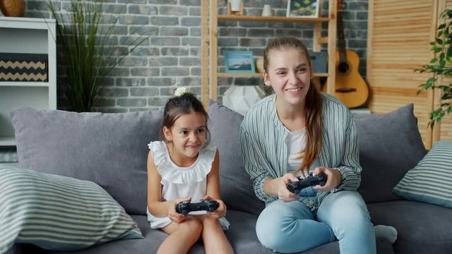 Mother and cute little daughter are playing video game at home having fun laughing together sitting on couch holding joysticks. People and lifestyle concept.