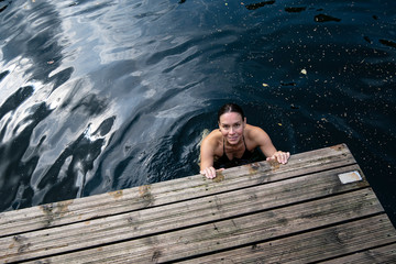 Smiling woman bathes in a clear lake, she holds her hands on the edge of a wooden pier and looks at the camera.