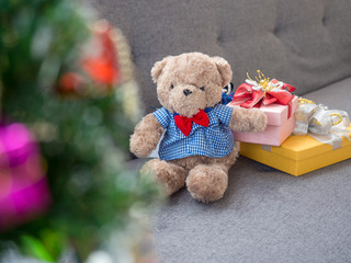 A cute little teddy bear and a gift box wrapped in red and yellow paper placed on a gray sofa.Foreground Christmas tree.