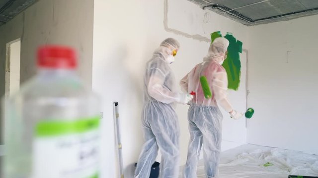 Dolly cam: positive young woman and guy in workwear and respirators dance lambada enter empty repairing room with white walls