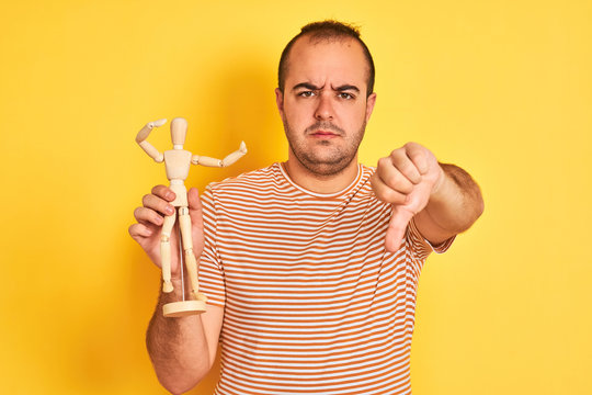Young man holding figure of art dummy standing over isolated yellow background with angry face, negative sign showing dislike with thumbs down, rejection concept - Powered by Adobe