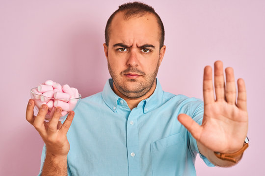 Young man holding bowl with marshmallows standing over isolated pink background with open hand doing stop sign with serious and confident expression, defense gesture