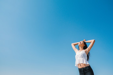 Happy woman with tanned slim body breathing fresh air raising her arms up, enjoying a sunny summer holiday on beach destination against blue sky, outdoors. Travel and well being lifestyle.