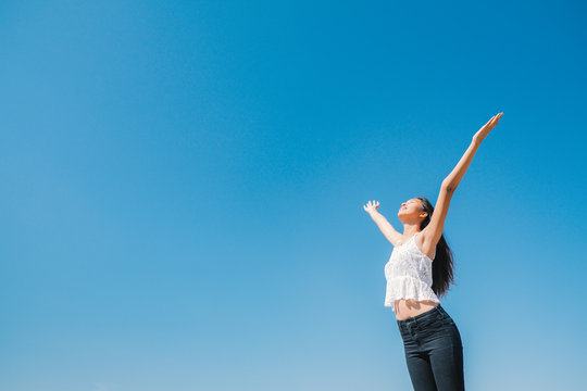 Happy woman with tanned slim body breathing fresh air raising her arms up, enjoying a sunny summer holiday on beach destination against blue sky, outdoors. Travel and well being lifestyle.