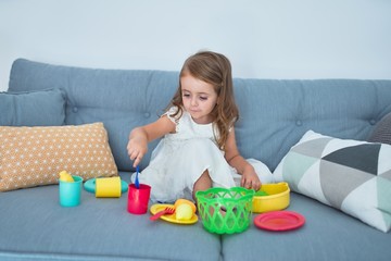 Adorable blonde toddler siting on the sofa cooking with food and cutlery toys at home