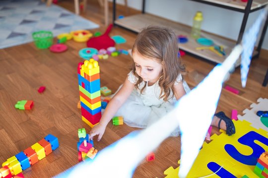 Adorable blonde toddler playing with building blocks around lots of toys at kindergarten
