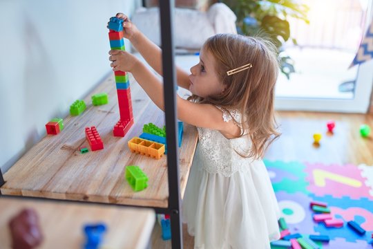 Adorable blonde toddler playing with building blocks around lots of toys at kindergarten