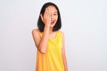 Young chinese woman wearing yellow casual t-shirt standing over isolated white background covering one eye with hand, confident smile on face and surprise emotion.