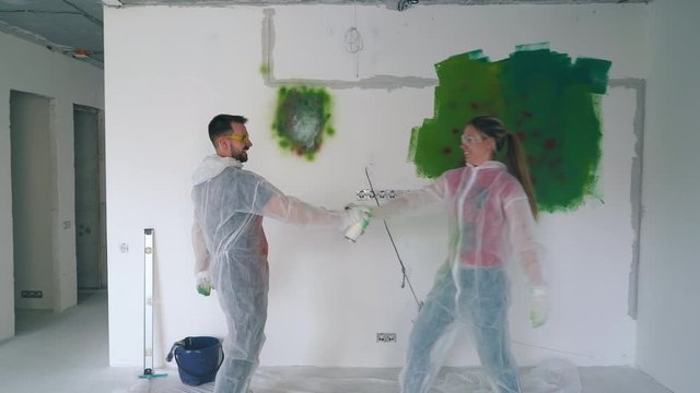 joyful guy and pretty girl in white protective suits fight with spray paints and kiss against room wall with color stains