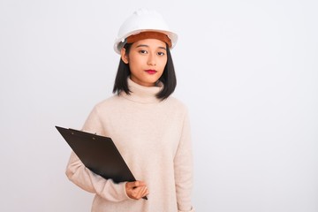 Chinese architect woman wearing helmet holding clipboard over isolated white background with a confident expression on smart face thinking serious