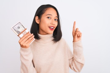 Young beautiful chinese woman holding dollars standing over isolated white background surprised with an idea or question pointing finger with happy face, number one