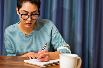 A woman wearing glasses sits at a table drinking coffee and writing in a journal