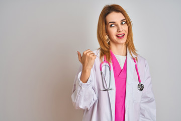 Redhead caucasian doctor woman wearing pink stethoscope over isolated background smiling with happy face looking and pointing to the side with thumb up.