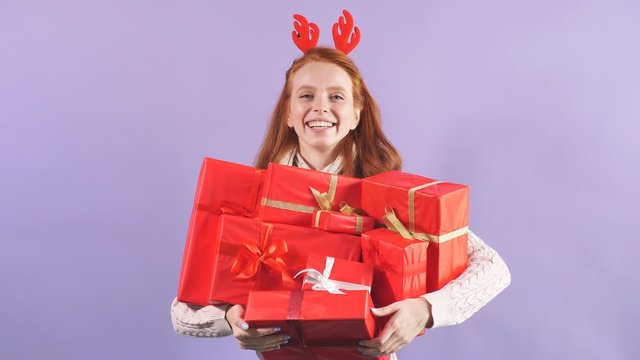 Smiling playful girl holding many beautiful Christmas present, finding lots of presents under Christmas tree, wearing funny red horns, looking at camera with cheerful face
