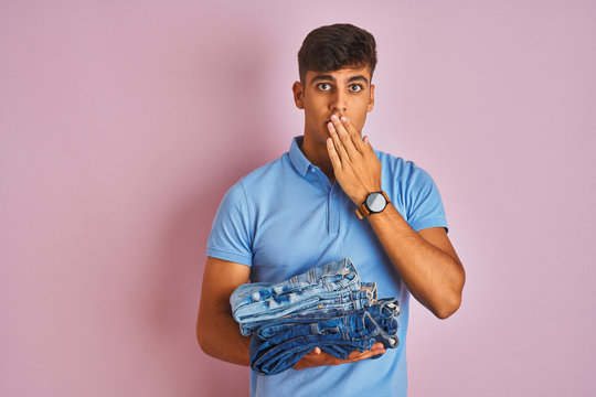 Young indian shopkeeper man holding folded jeans standing over isolated pink background cover mouth with hand shocked with shame for mistake, expression of fear, scared in silence, secret concept