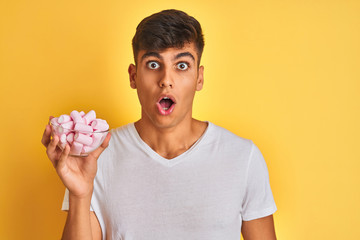 Young indian man holding bowl with marshmallows over isolated yellow background scared in shock with a surprise face, afraid and excited with fear expression