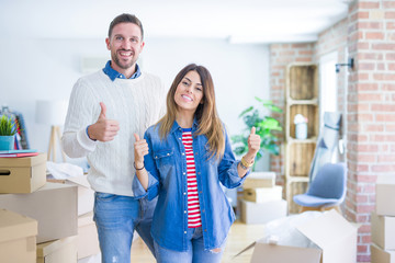 Young beautiful couple standing at new home around cardboard boxes success sign doing positive gesture with hand, thumbs up smiling and happy. Cheerful expression and winner gesture.