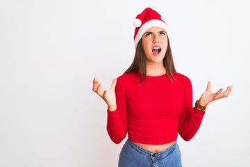 Young beautiful girl wearing Christmas Santa hat standing over isolated white background crazy and mad shouting and yelling with aggressive expression and arms raised. Frustration concept.
