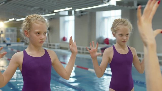 Panning of two female twins wearing similar purple swimming suits standing at swimming pool and repeating movements after young pretty woman standing in front of them