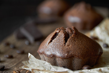 Homemade chocolate muffins on parchment with coffee beans and pieces of chocolate. Dark wooden background, closeup, selective focus