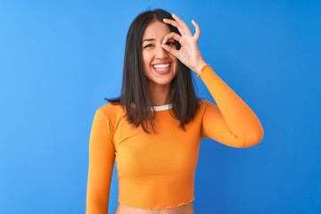 Young beautiful chinese woman wearing orange t-shirt standing over isolated blue background doing ok gesture with hand smiling, eye looking through fingers with happy face.