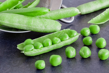geöffnete Erbsenschoten auf einem dunklen Tisch mit einem Sieb im Hintergrund