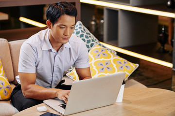 Young serious Vietnamese businessman spending work day in cafe, he is looking at computer screen...
