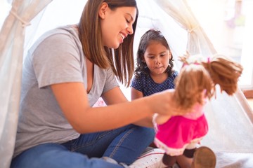 Beautiful teacher and toddler girl sitting on the floor playing with unicorn and doll inside tipi...