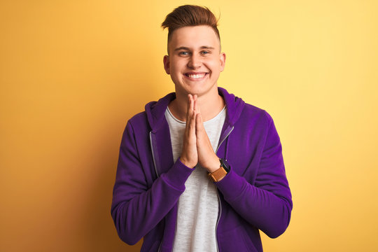 Young handsome man wearing purple sweatshirt standing over isolated yellow background praying with hands together asking for forgiveness smiling confident.