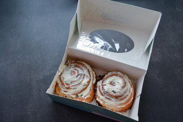 Two classic rolls with cinnamon and cheese cream on top in an open paper box  on a black, dark gray background